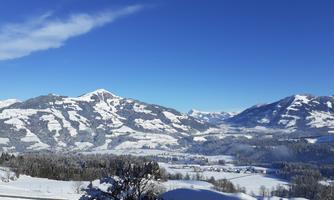 A winter landscape with snow-covered mountains and a clear blue sky. Below are snow-covered fields and trees.