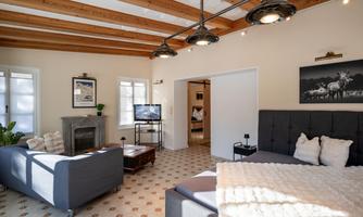 A modern living room with a wooden beam ceiling and light walls. There is a comfortable couch, a television, and a decorative fireplace.