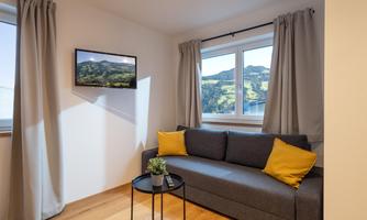 A cozy living space with a gray sofa and yellow cushions. There is a table and a television, as well as a window with a view of the mountains.