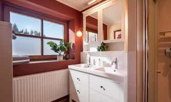 A modern bathroom with a white sink and two faucets. Large windows provide plenty of natural light, and green plants give the space a fresh touch.
