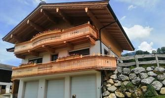 A beautiful, traditional chalet with wooden decorations and a balcony. In the background, green meadows and a clear sky can be seen.