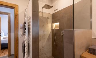 A modern shower in a bright bathroom with gray walls. In the background, a towel rack and a light wood shelf are visible.