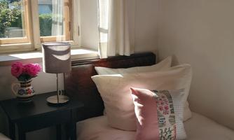 A cozy bedroom with two beds and a small lamp. The window lets in a lot of light and there are flowers on the nightstand.