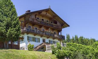 A traditional wooden house with balconies in a green setting. Trees and a blue sky can be seen in the background.