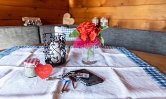 A table with a white tablecloth, on which stands a small vase with red flowers. Additionally, there are keys, a glass with a lid, and a candle in an artistic holder.