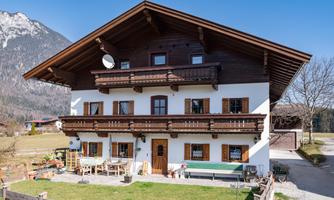 A traditional alpine house with several balconies and typical wooden details. In the background, mountains and a clear blue sky can be seen.