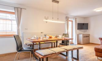A cozy dining area with a wooden table and benches. The room is brightly lit and features modern furniture as well as a television.