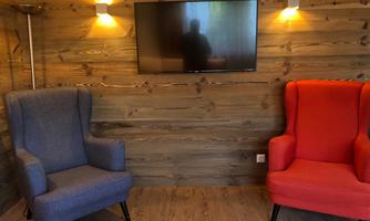 A cozy room with two armchairs, one blue and one red, in front of a wood-paneled wall. A television hangs on the wall, reflecting slightly.