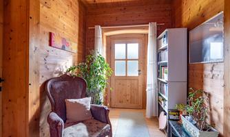 A cozy entrance area with wooden walls and a large window. On the left side, there is a comfortable armchair, and on the right, there is a shelf with books.