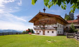 An old, traditional house in a rural setting with a green garden. The surrounding mountains are visible in the background, and the sky is clear.