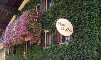 A charming building with green ivy and colorful flowers. The sign displays the name "Haus LUKAS."
