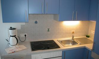 A modern kitchen with blue cabinets and a white countertop. It includes a kettle, a hot plate, and a sink.