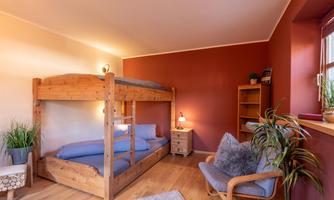 A cozy bedroom with a wooden bunk bed and a single bed. The walls are painted in a warm red hue and the room is decorated with plants and a shelf.