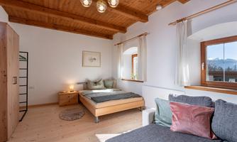 A bright bedroom with a wooden ceiling and large windows. There is a bed, a cozy armchair, and a nightstand.