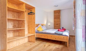 A cozy bedroom with a wooden shelf and a large bed. There are colorful cushions and a wooden floor.