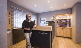 A friendly receptionist stands at the reception of House Lukas. The room is modern and inviting with wooden details and soft lighting.