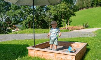 A small boy is standing in a sandbox under a sunshade. In the background, trees and a green meadow can be seen.