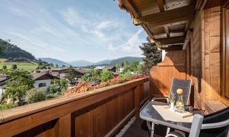A balcony with a view of a picturesque landscape and the mountains. A drink is on the table and flowers are blooming in the foreground.