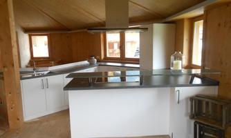 A modern kitchen with wood-paneled walls and large windows. There is a cooking island with a clear countertop and minimalist design.