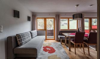 A modern living room with a cozy couch and a dining table. Large windows provide a view of the green surroundings.