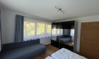 A cozy bedroom with a sofa, a mirrored wardrobe, and large windows. Bright walls and a wooden floor create an inviting atmosphere.