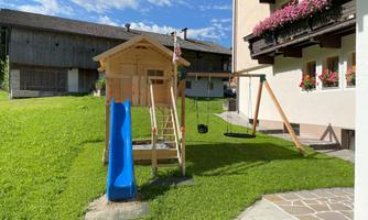 A playground with a slide, swing, and climbing frame. In the background, there is a garden and a house with flower windows.