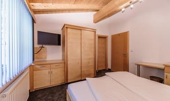 A modern bedroom with a double bed and wooden ceiling. There is a flat-screen TV and a desk next to a large wardrobe.