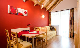 A cozy living kitchen with red walls and wooden ceilings. The table is set, and there's a view of a balcony with plenty of light.
