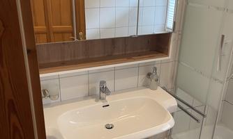 A modern bathroom with a sink, mirror, and a shower cabin. The walls are tiled in light colors, and there are wooden details.