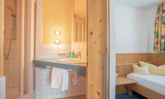 A modern bathroom with a shower and sink. In the background, a cozy bedroom with wooden details and colorful curtains is visible.