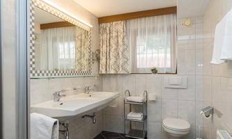 A modern bathroom with a large mirror and elegant lighting. There are gray tiles and simple sanitary facilities visible.