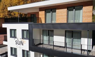 A modern building with large windows and balconies. The facade combines wood and white plaster, with the name "SUN" visible.