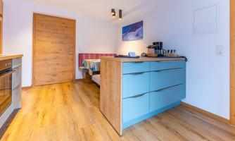 A modern kitchen with light blue cabinets and wooden flooring. In the background, a sofa and a wooden door can be seen.