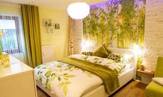 A modern bedroom with a bed in soft green tones. The walls are decorated with natural motifs and there is cozy lighting.