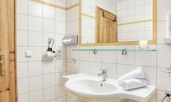 A simple bathroom with white tiles and a round sink. Towels are neatly arranged, and the warm wood of the door is reflected in the mirror.