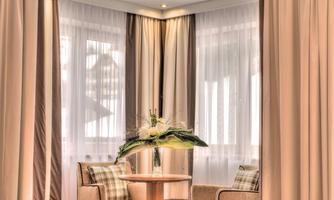 A cozy room with two chairs and a small table. The windows are adorned with light curtains and there is a beautiful flower arrangement.