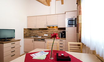 A modern kitchen unit with wooden details and a dining table. Next to a television, there is a vase with a flower on the table.