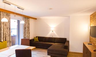 A modern living room with a comfortable sofa and a dining area. The walls are light-colored and decorated with elegant curtains.
