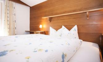 A cozy bedroom with a wooden design. The bed is adorned with floral bedding and there is soft light from a table lamp.