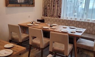 A cozy dining area with wooden tables and light chairs. A picture of a landscape hangs on the wall, and the light is bright and cheerful.