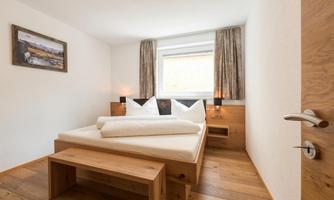 A cozy bedroom with a double bed and wooden floor. The room has large windows and a bright, friendly atmosphere.