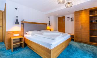 A modern bedroom with a large double bed and wooden details. The walls are light, and the floor is covered with carpet in a calm blue.