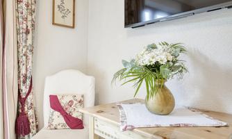 A cozy room with a white chair and a wooden table. On the table is a flower vase with fresh flowers and a embroidered cloth.