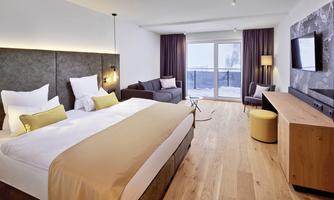 A modern hotel room with a large bed, a cozy sitting area, and an open balcony door. The floor is made of wood and the walls are in soft colors.