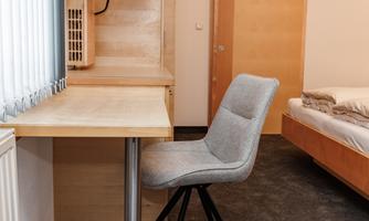 A simple room with a desk and a modern chair. In the background, a bed and a wooden wall are visible.