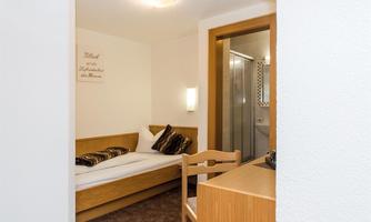 A simple room with a bed, a desk, and a chair. In the background, there is a bathroom with a shower.