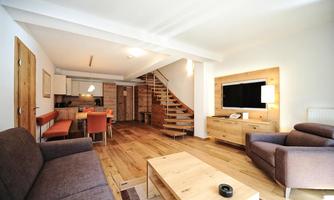 A modern living room with wooden flooring and an open kitchen. The furniture isSimple and comfortable, with a staircase in the background.