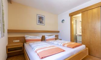 A cozy bedroom with two beds, orange bedding, and wooden paneling. A bathroom is visible in the background.