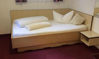 A simple room with a single bed, two pillows, and a white duvet. The room has carpet flooring and a window with curtains.