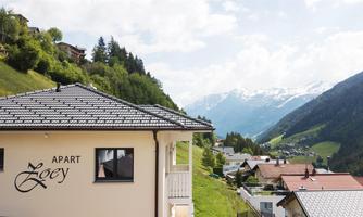 A modern apartment building called "Apart Zoey" in the mountains. In the background, the majestic Alps stretch out under a clear sky.